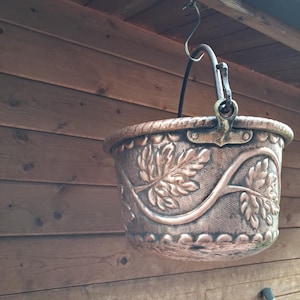 Op de afbeelding: Een koperkleurige, hangende ketel met reliëfblad- en wijnstokontwerpen. De ketel heeft een decoratieve rand en een handvat om op te hangen. De achtergrond is een houten muur.