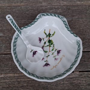 May include: White ceramic serving dish and spoon set. The dish has a scalloped edge with a green and white border. The dish and spoon feature a botanical illustration of purple flowers and green leaves. The text "Origanum vulgare" is printed on the dish.