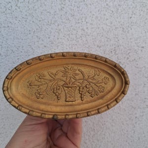 May include: An oval, gold-coloured decorative tray with a raised floral design. The tray features a central vase with flowers and grapes, surrounded by a beaded border. The tray is held in a hand against a neutral background.