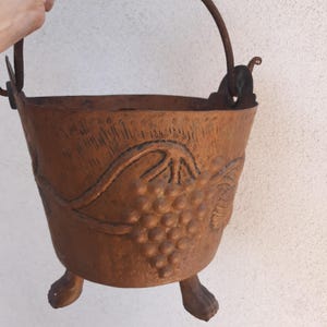 Puede incluir: Un cubo de metal marrón rústico con un diseño de vid y tres patas. El cubo tiene un asa con un gancho en el extremo.