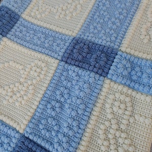 Puede incluir: Una manta de ganchillo hecha a mano con un diseño de patchwork. La manta presenta cuadrados en varios tonos de azul y crema, con patrones texturizados que incluyen bolitas y puntadas de canalé. La estética general es acogedora y artesanal.