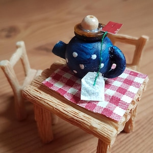 Könnte beinhalten: Eine blaue Miniatur-Teekanne mit weißen Punkten und einem Holzdeckel, die auf einer rot-weiß karierten Tischdecke auf einem Holztisch steht.