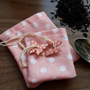 Könnte beinhalten: Zwei rosa Stoffbeutel mit weißen Punkten und Kordelzug. Die Beutel sind übereinander gestapelt. Jeder Beutel hat eine Holzschmetterlingsverzierung mit rosa und weißen Blumendetails.