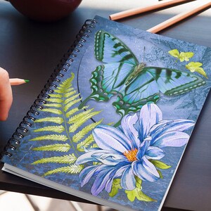 Könnte beinhalten: Ein blaues Spiralnotizbuch mit einem blauen Schmetterling, einem grünen Farn und einer lila Blume auf dem Einband.
