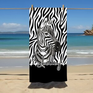 Könnte beinhalten: Schwarz-weißes Zebra-Muster-Strandtuch mit einem schwarz-weißen Zebra-Kopf-Motiv und dem Namen "Shelley" in schwarzer Kursivschrift.