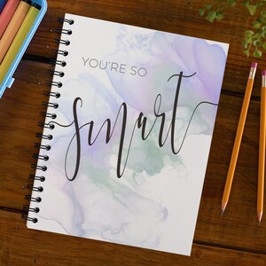 Op de afbeelding: Een spiraalgebonden notitieboek met een aquarel achtergrond in tinten van paars, groen en blauw. De tekst "YOU'RE SO SMART" is in zwarte cursieve letters op de voorkant geschreven.