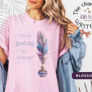 Könnte beinhalten: Hellrosa T-Shirt mit dem Text "It's a good day for writing" und einer Feder-Illustration. Das Shirt hat auch den Text "The charming Gift for WRITERS" und das Wort "BLOSSOM".