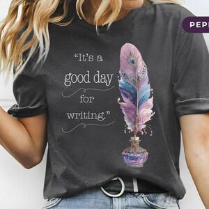 Könnte beinhalten: Dunkelgraues T-Shirt mit dem Text "It's a good day for writing." Das Shirt zeigt ein farbenfrohes Federdesign in Lila, Blau und Rosa sowie ein dekoratives Tintenfass.
