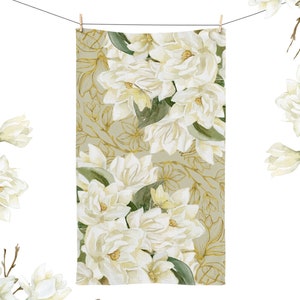 May include: A white floral patterned towel hanging from a clothesline. The towel features a repeating pattern of white magnolia flowers and green leaves against a light green background. Gold-colored floral outlines add detail. The towel is held by two wooden clothespins.