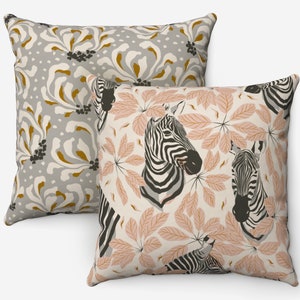 May include: Two decorative throw pillows. One pillow features a gray background with a floral pattern in white, gold, and black. The other pillow has a cream background with zebra heads and pink leaves. The pillows are square.