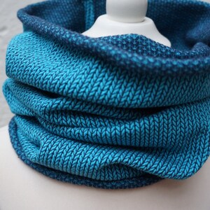 Könnte beinhalten: Ein teal- und marineblauer Strickschal. Der Schal besteht aus einem weichen, strukturierten Stoff mit einem sich wiederholenden Muster. Der Schal wird auf einem weißen Schaufensterpuppenkopf präsentiert. Ein vielseitiges Accessoire für kaltes Wetter.
