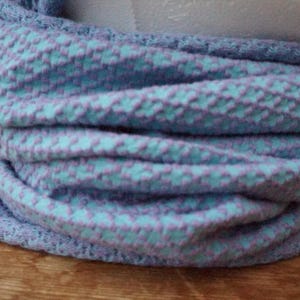 Könnte beinhalten: Ein weicher, gestrickter Rundschal in Blau- und Lavendeltönen. Der Stoff hat ein strukturiertes, gewebtes Muster mit einem sich wiederholenden Design aus kleinen Quadraten. Der Schal ist um eine weiße Form gewickelt.