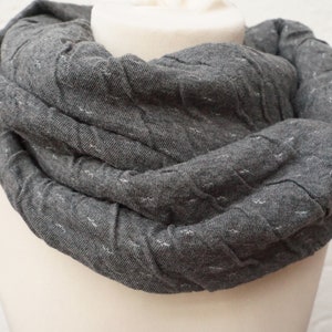 Könnte beinhalten: Grauer Loop-Schal aus gestepptem Stoff, auf einer Schaufensterpuppe präsentiert. Der Schal ist dunkelgrau mit kleinen weißen Sprenkeln im gesamten Material. Der Stoff ist in einer kreisförmigen Form gefaltet.