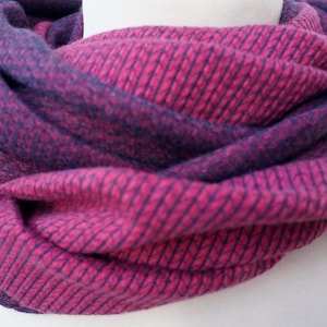Könnte beinhalten: Ein gestrickter Loop-Schal in Rosa- und Lilatönen. Der Stoff hat ein strukturiertes Muster. Der Schal wird in einer kreisförmigen Form präsentiert, die sein Design hervorhebt. Dieses Accessoire ist ideal, um Wärme und Stil zu verleihen.
