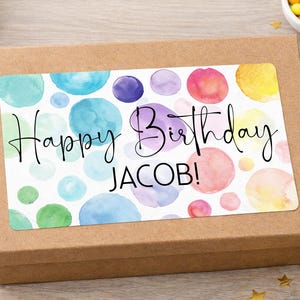 May include: A brown gift box with a watercolour-style label. The label features colourful circles and the text "Happy Birthday JACOB!" in a handwritten font. Gold stars and a small bowl of sweets are nearby.
