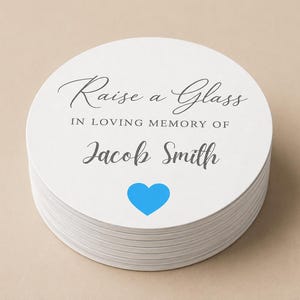 May include: A pile of white, circular coasters. The text reads "Raise a Glass in Loving Memory of Jacob Smith" in a script font, with a blue heart beneath the name. These coasters are for a memorial.