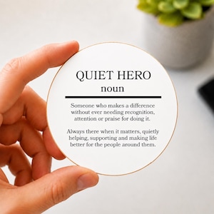 May include: A round white coaster with a thin gold rim featuring black text that reads: QUIET HERO noun. Someone who makes a difference without ever needing recognition, attention or praise for doing it. Always there when it matters, quietly helping.