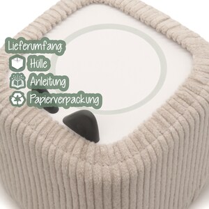 May include: A close-up of a beige, corduroy-textured, square-shaped object with a white circular design on top. The object has two black, triangular-shaped features. The text "Lieferumfang: H&uuml;lle, Anleitung, Papierverpackung" is visible.