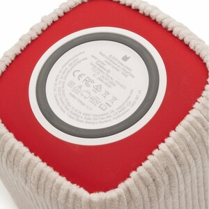 May include: Close-up of a red and beige cube-shaped speaker. The bottom of the speaker has a white circular label with text and symbols. The speaker has a corduroy-like texture around the edges.