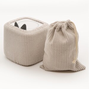 May include: A beige corduroy cube-shaped object with black ear-like protrusions, next to a matching drawstring bag. The cube has a circular opening on top. The bag is closed with a beige drawstring.