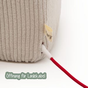 May include: Close-up of a beige, corduroy-textured object with a white charging cable and a red cord. A small tag with the word "Mary" is attached. The text "&Ouml;ffnung f&uuml;r Ladekabel" is at the bottom.