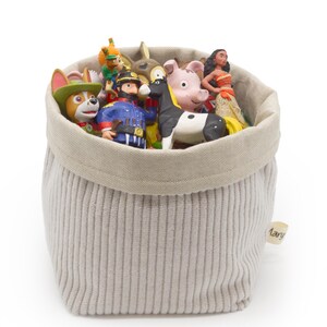May include: A gray corduroy storage basket filled with colorful toy figurines. The toys include a cartoon dog, a pig, a black and white horse, and a doll. The basket has a neutral-colored rim and a small tag that reads "Mary".