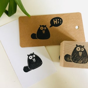 Könnte beinhalten: Eine schwarze Biber-Illustration ist auf einem Kraftpapier-Etikett, einer weißen Karte und einem Holzstempel abgebildet. Das Etikett hat eine Sprechblase mit "Hi!" darin. Der Biber hat große Augen und Schneidezähne.