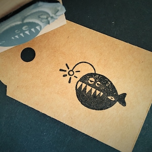 Puede incluir: Una etiqueta de papel marrón con un sello negro de pez rape de dibujos animados. El sello presenta un pez con dientes afilados y un señuelo brillante. Un sello a juego es parcialmente visible encima de la etiqueta.
