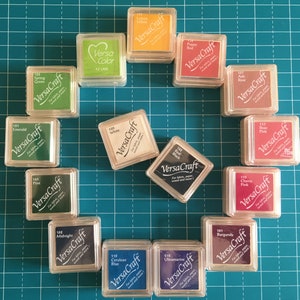 May include: A collection of VersaCraft ink pads in various colors, including green, yellow, red, and blue. Each pad is in a clear plastic case with the color name and brand printed on the label. The ink pads are arranged on a teal cutting mat.