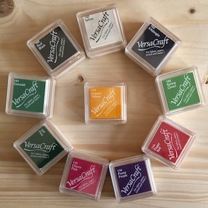 May include: A collection of VersaCraft ink pads in various colors, including Real Black, White, Midnight, Emerald, Lemon Yellow, Spring Green, Pine, Cherry Pink, Peony Purple, and Poppy Red. Each pad is in a clear plastic case, arranged on a light wood surface.