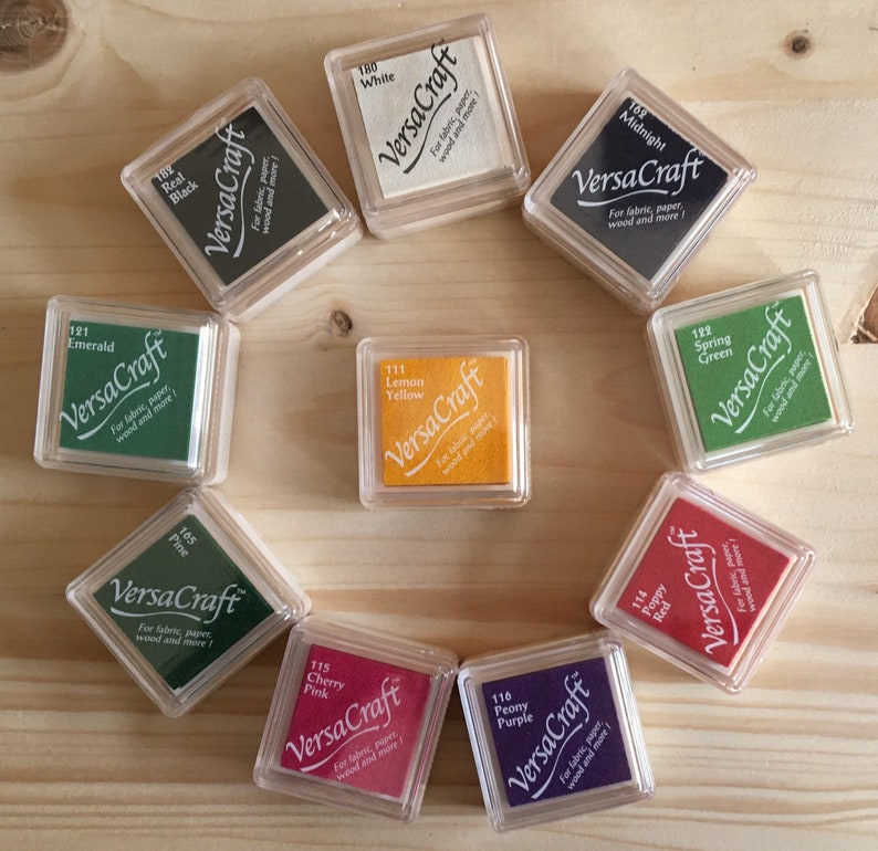 May include: A collection of VersaCraft ink pads in various colors, including Real Black, White, Midnight, Emerald, Lemon Yellow, Spring Green, Pine, Cherry Pink, Peony Purple, and Poppy Red. Each pad is in a clear plastic case, arranged on a light wood surface.