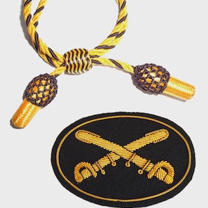 Könnte beinhalten: Ein dekoratives Militär-Lanyard mit gelb-schwarzem gedrehtem Kordel, einem zentralen Knoten und zwei dekorativen Quasten. Darunter ein ovales Abzeichen mit gekreuzten Schwertern, die auf schwarzem Grund mit Goldfaden gestickt sind.
