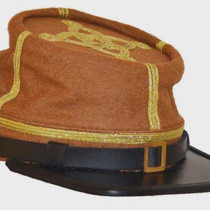 Może przedstawiać: Brązowy kapelusz w stylu wojskowym ze złotymi wykończeniami i akcentami. Kapelusz posiada czarny rondo i czarny skórzany pasek ze złotą klamrą. Złote elementy dekoracyjne znajdują się na górze kapelusza.