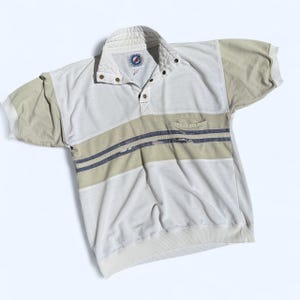 May include: A short-sleeve polo shirt featuring a white base with beige sleeves and a horizontal beige chest stripe accented by two thin dark blue lines. The shirt includes a quilted collar, a three-button placket, and a small chest pocket.