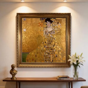 Op de afbeelding: Een ingelijst schilderij van een vrouw in een gouden jurk met ingewikkelde geometrische patronen, tentoongesteld aan een muur boven een houten bank. Het kunstwerk heeft een opvallende achtergrond van bladgoud, donker haar en een versierd lijfje.