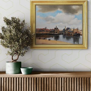 a painting of a cityscape with a river in the foreground, hanging on a wall above a wooden shelf with a potted plant and a small bowl on it.