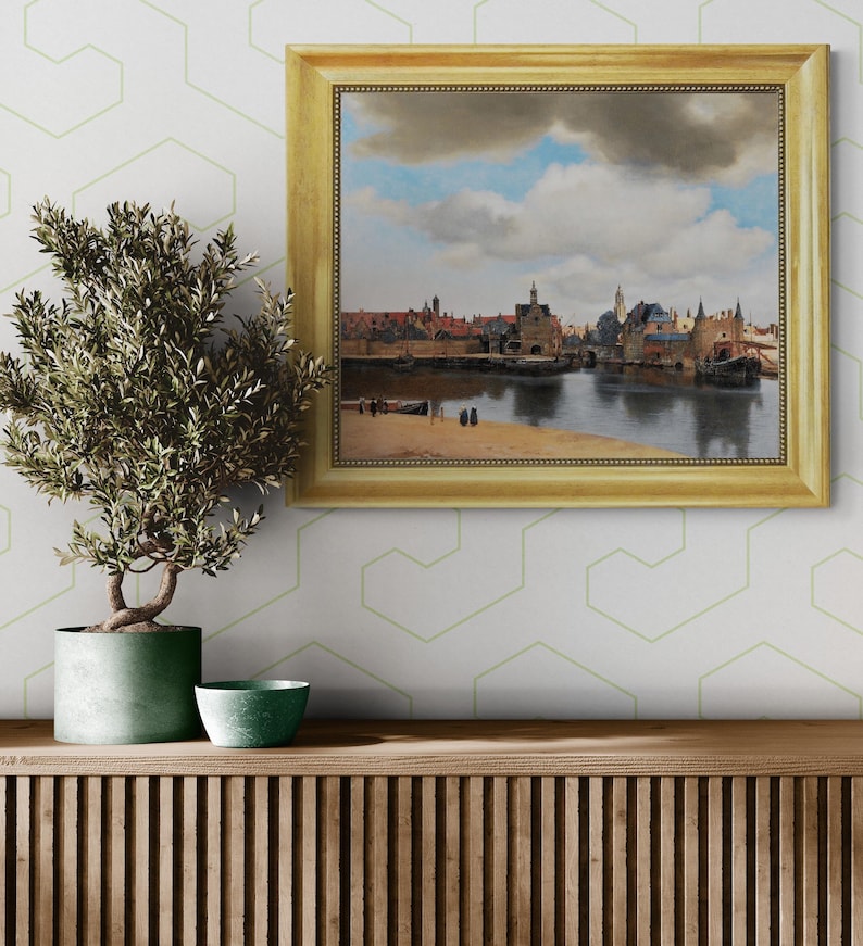 a painting of a cityscape with a river in the foreground, hanging on a wall above a wooden shelf with a potted plant and a small bowl on it.