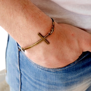 Könnte beinhalten: Ein schwarzes Armband mit einem goldfarbenen Kreuzanhänger. Das Armband wird am Handgelenk getragen.