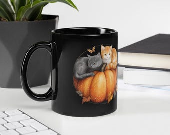 May include: Black ceramic mug featuring a watercolor illustration of a gray cat and an orange tabby cat resting on a pumpkin. A small butterfly and autumn leaves are also depicted. The mug has a black handle.