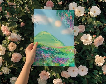 May include: A hand holds a print of a green hill with purple flowers in the foreground and a blue sky with white clouds in the background.