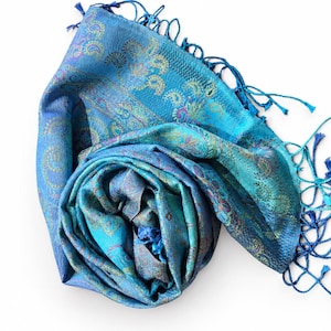 May include: A vibrant blue scarf featuring an intricate paisley pattern in shades of teal, purple, and gold. The fabric has a subtle sheen and is finished with long, delicate blue fringe tassels along the edges, creating a textured appearance.