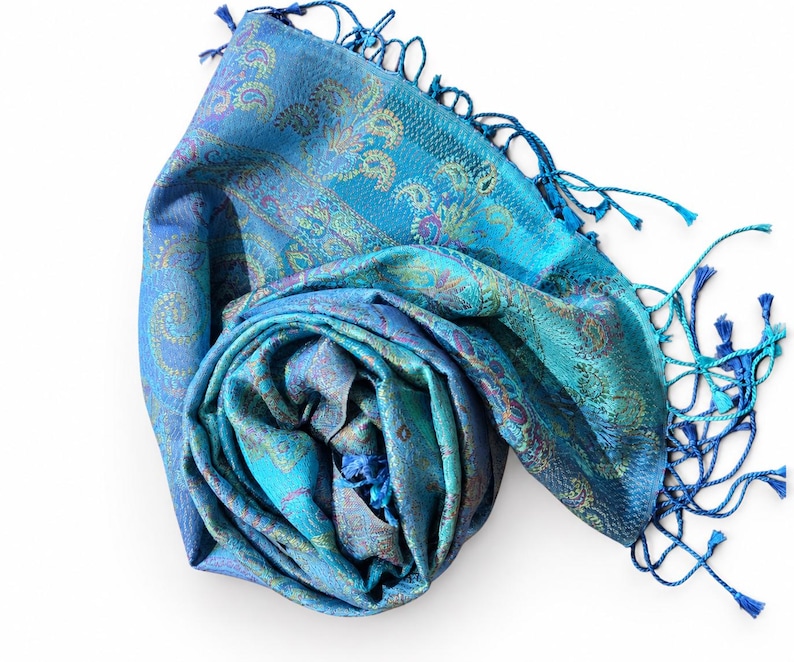 May include: A vibrant blue scarf featuring an intricate paisley pattern in shades of teal, purple, and gold. The fabric has a subtle sheen and is finished with long, delicate blue fringe tassels along the edges, creating a textured appearance.
