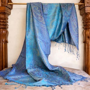 May include: A rectangular scarf featuring a vibrant blue and teal paisley pattern with intricate swirling designs. The fabric has a subtle sheen and is finished with delicate fringe detailing along the edges, creating a textured appearance.
