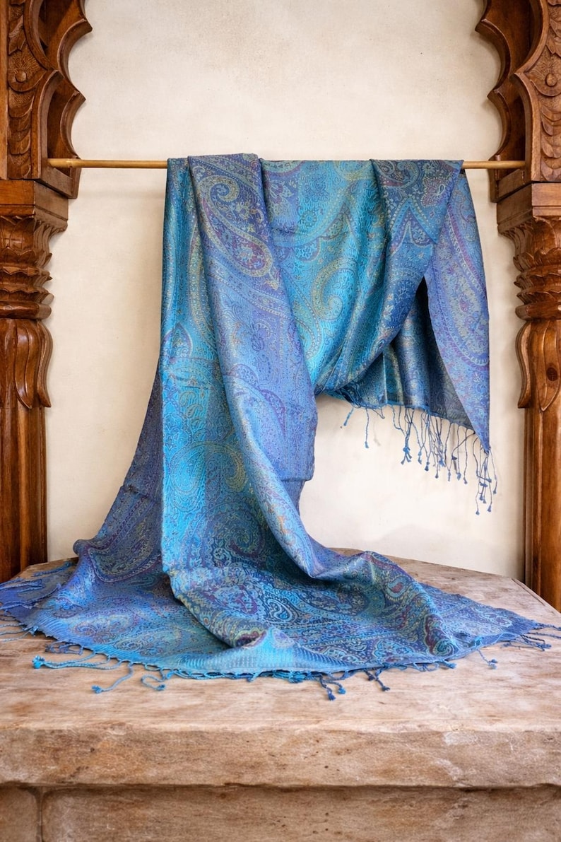 May include: A rectangular scarf featuring a vibrant blue and teal paisley pattern with intricate swirling designs. The fabric has a subtle sheen and is finished with delicate fringe detailing along the edges, creating a textured appearance.