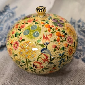 May include: A round, cream-coloured box with a floral design. The box is decorated with a variety of colourful flowers, including pink, yellow, blue, and orange. The box has a lid with a small knob on top.