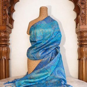 May include: A vibrant blue shawl featuring an intricate, shimmering paisley pattern in shades of teal, gold, and deep azure. The fabric has a subtle metallic sheen and is finished with delicate, matching blue fringe along the bottom edge.
