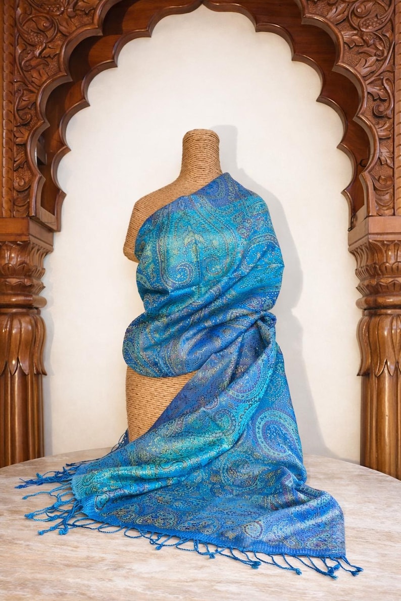 May include: A vibrant blue shawl featuring an intricate, shimmering paisley pattern in shades of teal, gold, and deep azure. The fabric has a subtle metallic sheen and is finished with delicate, matching blue fringe along the bottom edge.