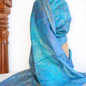 May include: A rectangular scarf featuring a vibrant turquoise and blue paisley pattern with intricate woven details. The fabric displays a subtle sheen and is finished with matching blue fringe along the bottom edge, creating a textured appearance.