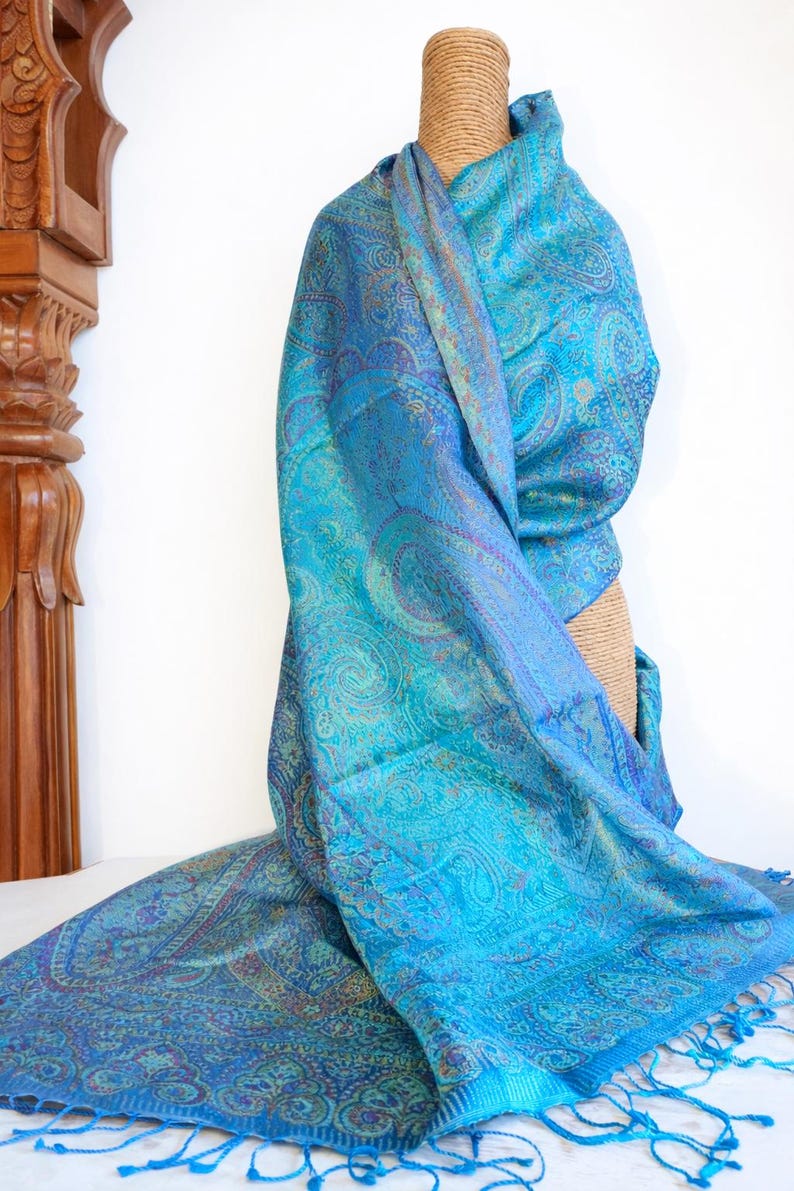 May include: A rectangular scarf featuring a vibrant turquoise and blue paisley pattern with intricate woven details. The fabric displays a subtle sheen and is finished with matching blue fringe along the bottom edge, creating a textured appearance.