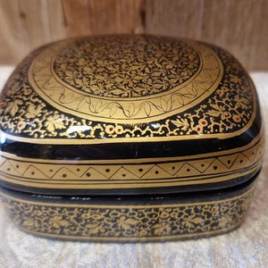Pode incluir: Uma caixa quadrada preta com intrincados desenhos florais dourados e uma borda dourada. A caixa é feita de papel machê e é um artesanato tradicional indiano.